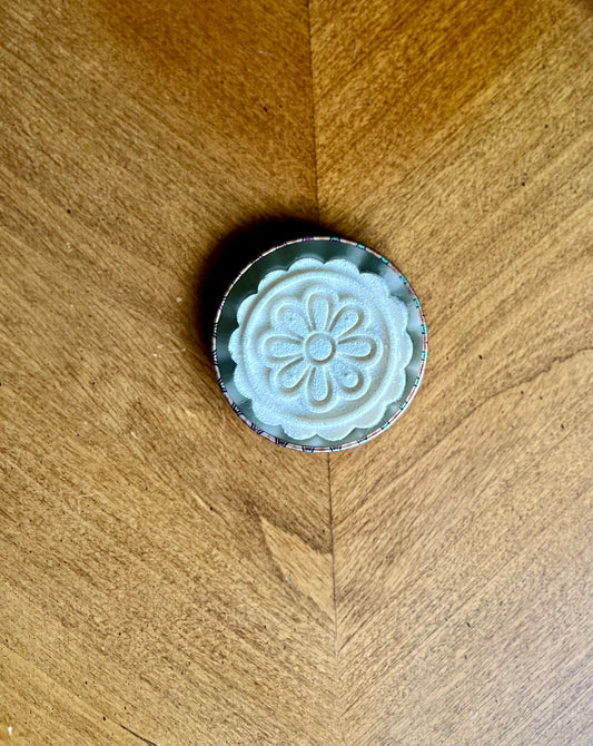 A single shower steamer stamped to look like a flower sits on a lid on a warmly lit wooden table.